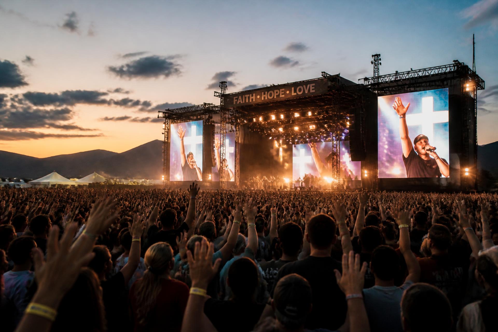 Crowd with hands raised in worship facing a golden-lit main stage at dusk with the Nevada mountains behind